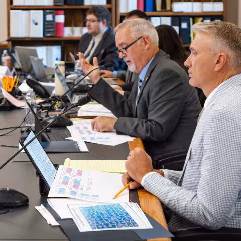man seated at a table with a computer and microphone, surrounded by paperwork and others