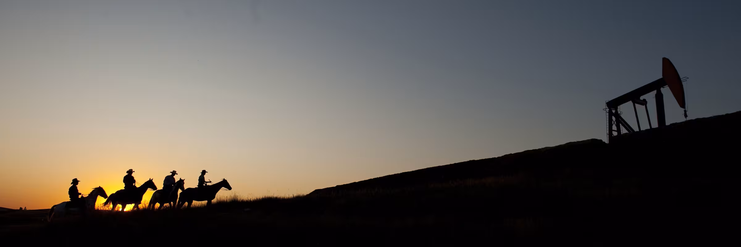 Dark silhouette of men on horseback approaching an oil derrick in front of a dakota unset